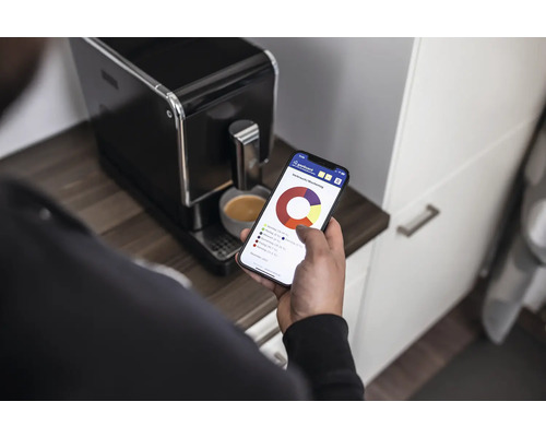 Eine Person betrachtet auf einem Smartphone eine Energieverbrauchsübersicht neben einer Kaffeemaschine.