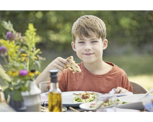Junge isst an einem gedeckten Tisch im Freien.