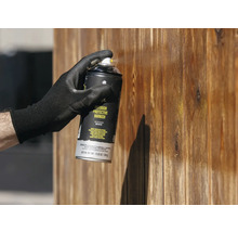 Außen-Schutzlack wird mit Handschuhen auf eine Holzwand gesprüht.