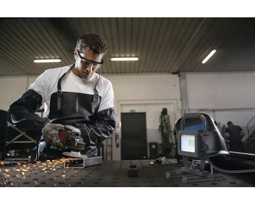 Ein Mann mit Schutzbrille, Schürze und Handschuhen schleift Metall mit einer Flex, während ein Baustrahler im Vordergrund steht.