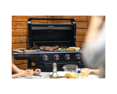 Schwarzer Gasgrill mit Grillgut und Gemüse auf dem Rost vor Holzwand