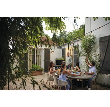 Gartenszene mit Grill, Tisch und sitzenden Personen unter einer Lichterkette