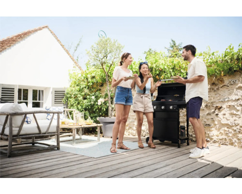Drei Personen grillen auf einer Terrasse mit einem Gasgrill, umgeben von Gartenmöbeln und Pflanzen.