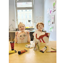 Zwei Kinder präsentieren bemalte Bilder, Farben und Pinsel liegen auf dem Tisch.