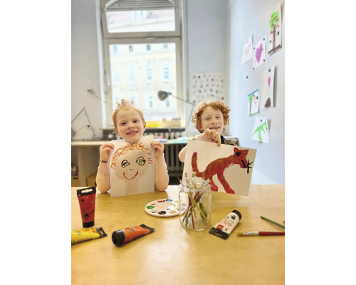 Zwei Kinder zeigen ihre mit Farbe gemalten Bilder in einem hellen Raum.