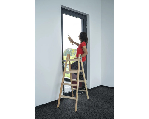 Frau steht auf einer Holzleiter und reinigt eine Fensterscheibe in einem Raum.