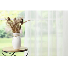 Dekorative Trockenblumen in weißer Vase auf kleinem Tisch vor einem Fenster mit Vorhang.