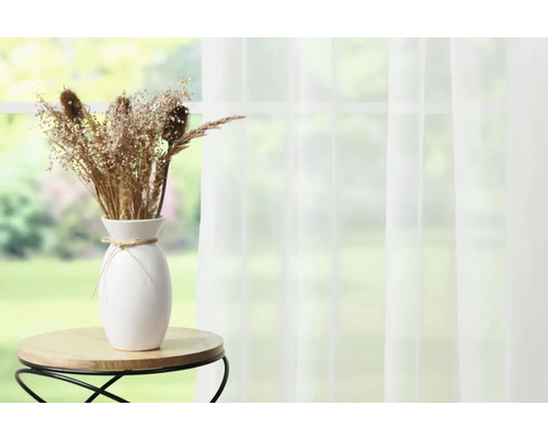 Dekorative Trockenblumen in weißer Vase auf kleinem Tisch vor einem Fenster mit Vorhang.