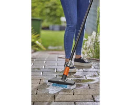 Eine Person reinigt mit einer Bürste und Stiel Pflastersteine im Freien.