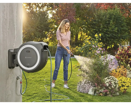 Frau bewässert Blumen mit einer an der Wand montierten Schlauchtrommel im Garten.