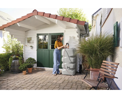 Frau befüllt einen grauen Regenwassertank auf einer Terrasse vor einem Gartenhaus.