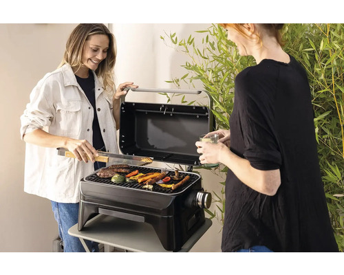 Zwei Frauen grillen Gemüse und Fleisch auf einem tragbaren Grill im Freien.