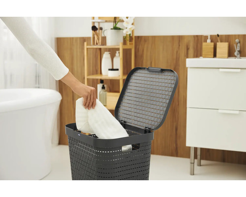 Szene im Badezimmer mit einem Wäschekorb mit Deckel, in den eine Person ein weißes Handtuch legt.