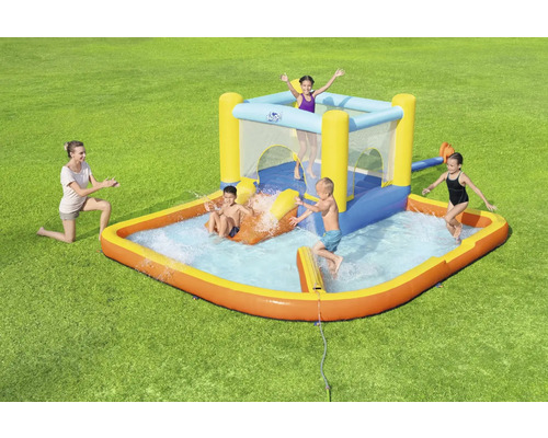 Aufblasbares Kinderplanschbecken mit Rutsche und Hüpfburg auf einer grünen Wiese mit spielenden Kindern und einer Frau.
