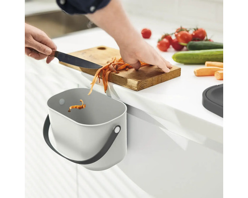 Küchenszene mit Person, Schneidebrett, Gemüseresten und einem an der Küchenschranktür befestigten Abfallbehälter.