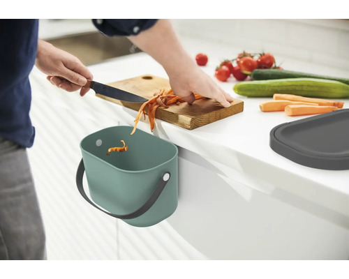 Eine Person schneidet Gemüse auf einem Holzbrett über einem Komposteimer in der Küche.