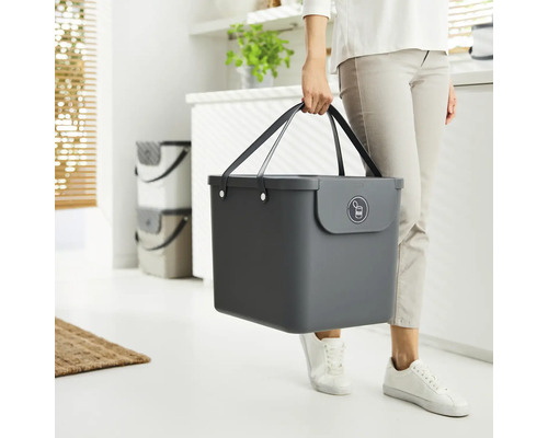 Eine Person trägt einen grauen Abfallbehälter mit Griffen und einem Symbol für Metallrecycling.