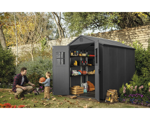 Gartenszene mit einem Gerätehaus aus Kunststoff mit geöffneten Türen, Regalen und Lagergegenständen, ein Mann und ein Kind spielen mit einem Ball