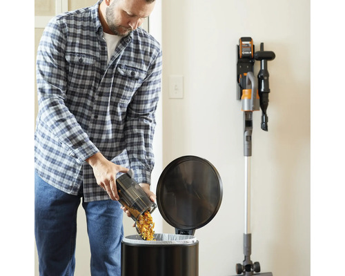 Ein Mann leert den Staubbehälter eines Akku-Staubsaugers in einen Mülleimer. An der Wand befindet sich eine Ladestation mit einem Staubsauger.