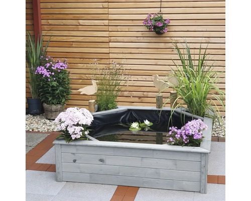 Gartenszene mit einem erhöhten Teich, umgeben von Blumen und Pflanzen vor einem Holzzaun.