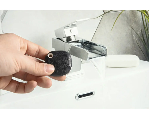 Hand hält einen schwarzen, quadratischen Wasserdetektor vor einem Waschbecken mit Wasserhahn