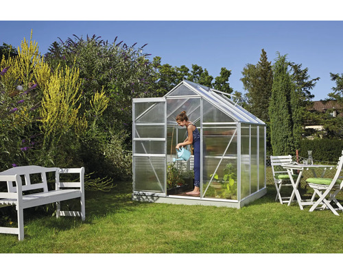 Gewächshaus im Garten mit einer Frau, die Pflanzen gießt, sowie einer Sitzgruppe und einer Bank.