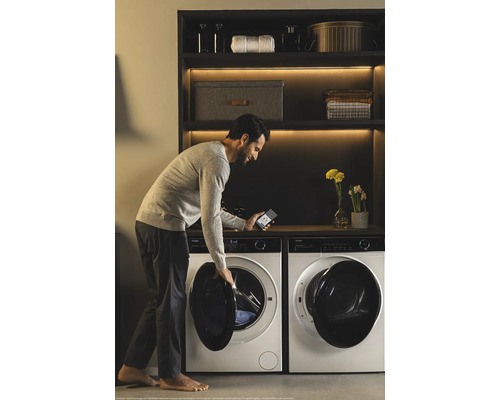 Mann bedient Waschmaschine mit Smartphone App neben Trockner und Regal