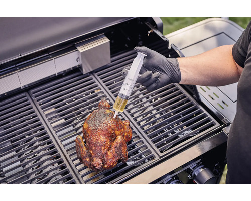 Eine Person injiziert mit einer Spritze Flüssigkeit in ein gegrilltes Hähnchen auf einem Grill.