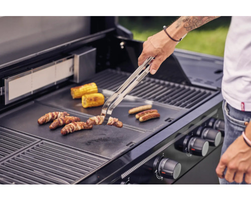 Person grillt Würstchen, Speck und Maiskolben auf einem Grill.