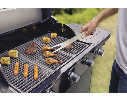 Ein Grill mit Grillgut wie Steaks, Würstchen, Hähnchen und Maiskolben wird mit einer Grillzange gewendet.