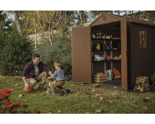 Szene mit einem Kunststoff-Gartengerätehaus mit Regalen, das mit verschiedenen Gegenständen gefüllt ist, während ein Mann und ein Kind auf dem Rasen spielen.