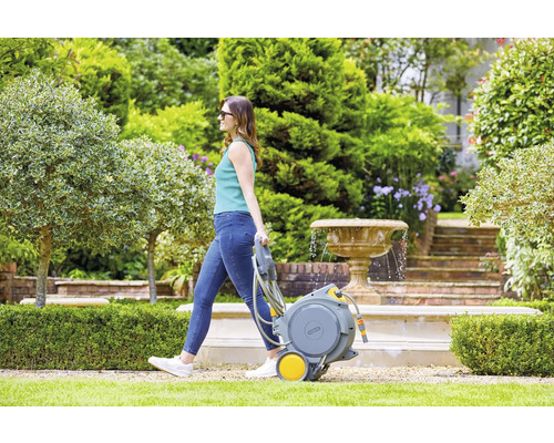 Frau zieht einen Gartenschlauchwagen in einem Garten mit Springbrunnen.