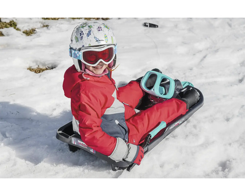 Ein Kind mit Helm und Skibrille sitzt auf einem Schlitten im Schnee.