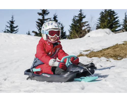 Kind mit Helm und Skibrille auf einem Rodelschlitten im Schnee.