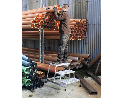 Eine Person auf einer Trittleiter hantiert mit orangefarbenen Rohren in einem Lager.