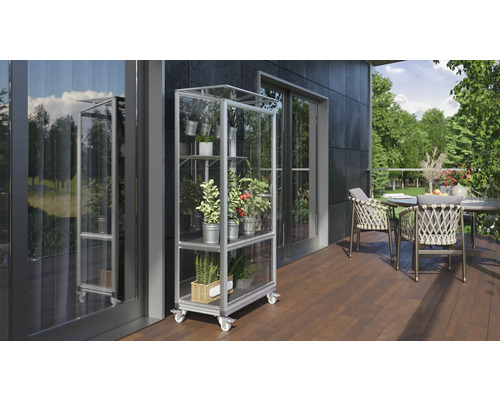 Eine Szene im Freien mit einem Pflanzenregal auf Rollen, das mit verschiedenen Pflanzen in Töpfen gefüllt ist und auf einer Terrasse neben einem Tisch mit Stühlen steht.