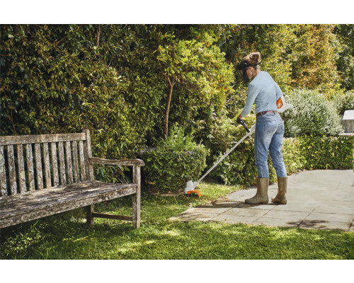 Frau arbeitet mit einem Rasentrimmer im Garten, neben einer Holzbank.