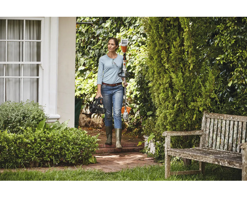 Frau trägt Akku-Motorsense und Akku-Heckenschere in gepflegtem Garten