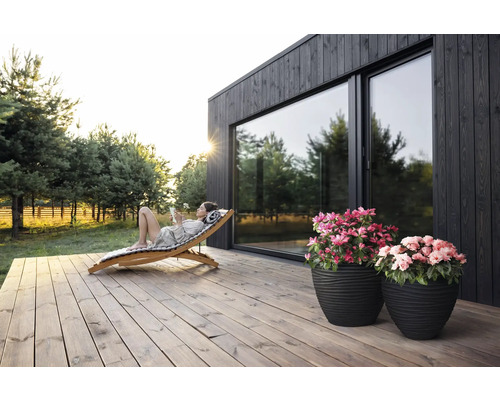 "Szene mit Frau, die sich auf einer Holzliege auf einer Holzterrasse vor einem Haus mit großen Fenstern entspannt