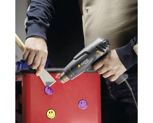 Eine Person entfernt mit einer Heißluftpistole und einem Spachtel Aufkleber von einer Oberfläche.