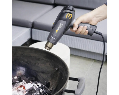 Eine Person zündet mit einem Heißluftfön Holzkohle in einem Grill an.