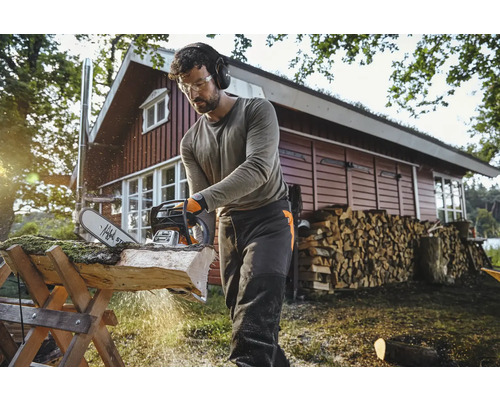 Mann mit Schutzbrille und Gehörschutz sägt Holz mit einer Motorsäge auf einem Holzbock im Freien.