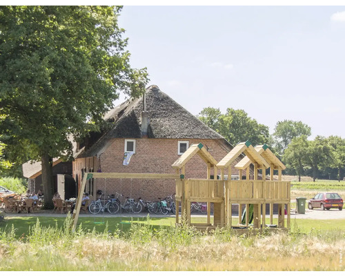 Spielhaus mit Stelzen P8S Douglasie HY-Land 680 x 920 cm Holz 2 Brettschaukeln, Abdeckplane, Schaukel, Sandkasten, Rutsche, Dacheindeckung, Grundturm, Kletterwand, Klettersteine Holzspielplatz mit Schaukel und Rutsche vor Backsteingebäude mit Strohdach