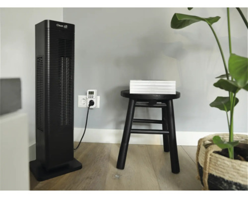 Raumszene mit einem Clean Air Turmventilator, einer Zeitschaltuhr, einem Hocker mit Büchern und einer Pflanze.