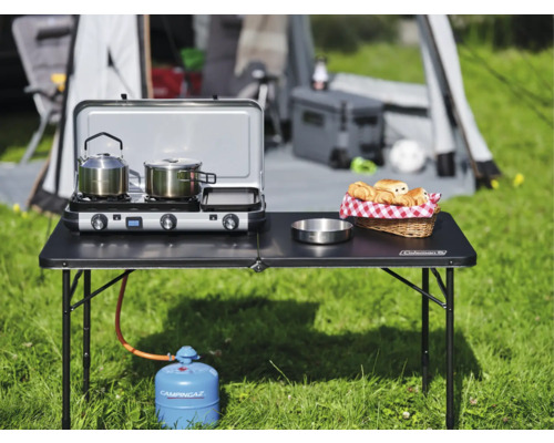 Campingszene mit Tisch, Gaskocher, Kochutensilien und Lebensmitteln vor Zelt