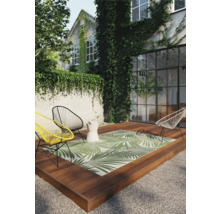 Terrasse mit Outdoor Teppich mit Palmenblattmuster, Stühlen und Beistelltisch vor einem Haus mit Pflanzenbewuchs.