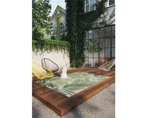 Terrasse mit Outdoor Teppich mit Palmenblattmuster, Stühlen und Beistelltisch vor einem Haus mit Pflanzenbewuchs.
