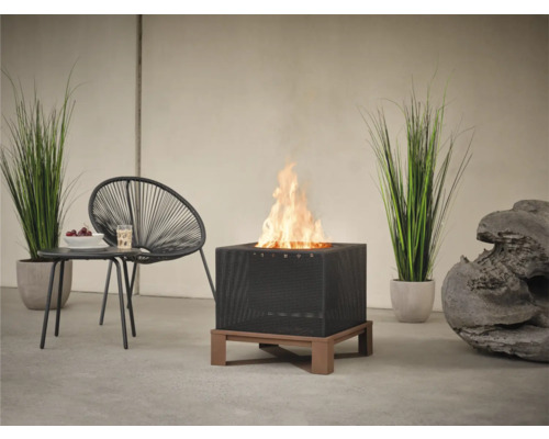 Quadratische Feuerstelle mit Flammen, Rattan-Design und Holzfuß auf einer Terrasse, ergänzt durch einen Stuhl und einen Tisch.