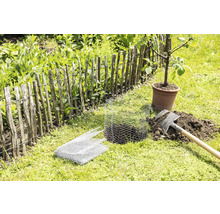 Gartenszene mit einem Holzzaun, einem jungen Baum im Topf, einem Spaten und einem Drahtgeflecht zum Schutz von Pflanzen.