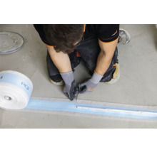 Handwerker schneidet Dichtband mit einem Messer auf einem Betonboden zu. Rolle des Dichtbands und ein Messwerkzeug sind auch sichtbar.
